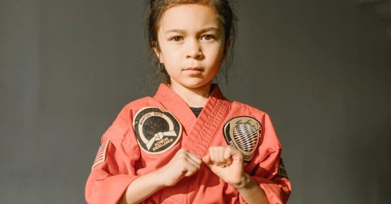A determined young girl wearing a red martial arts uniform with patches, standing in a ready stance to showcase preschool martial arts benefits such as confidence and self-esteem.